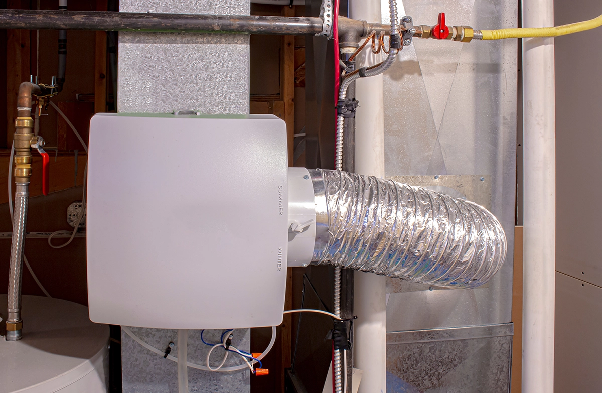 Whole-house humidifier mounted to ductwork with flexible bypass duct connected to a forced-air HVAC system. Best for blogs about whole-house humidifiers, dry indoor air, winter humidity control, and HVAC humidifier installation or maintenance.