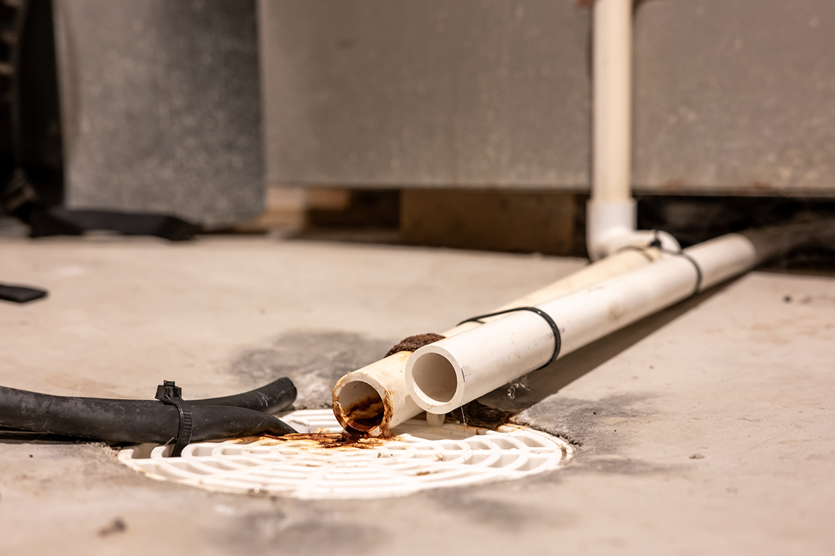 PVC drain lines discharging near a floor drain with visible rust staining on the concrete surface. Best for blogs about condensate drain line problems, HVAC drain line clog, AC leaking water, furnace leaking water, why is my HVAC dripping water, and condensate line maintenance.