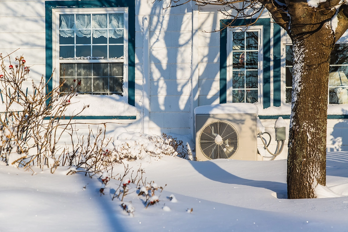 Outdoor heat pump unit installed beside a home in snowy winter conditions. Best for blogs about heat pumps in winter, should a heat pump run in snow, winter HVAC performance, cold-weather heating, outdoor unit in snow, and heat pump defrost cycles.