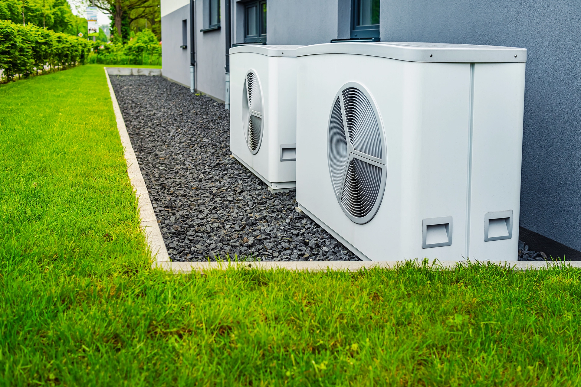 Two outdoor heat pump condenser units installed along the exterior wall of a home. Best for blogs about heat pump installation, heat pump vs AC, new HVAC system cost/value, and energy-efficient home cooling systems.